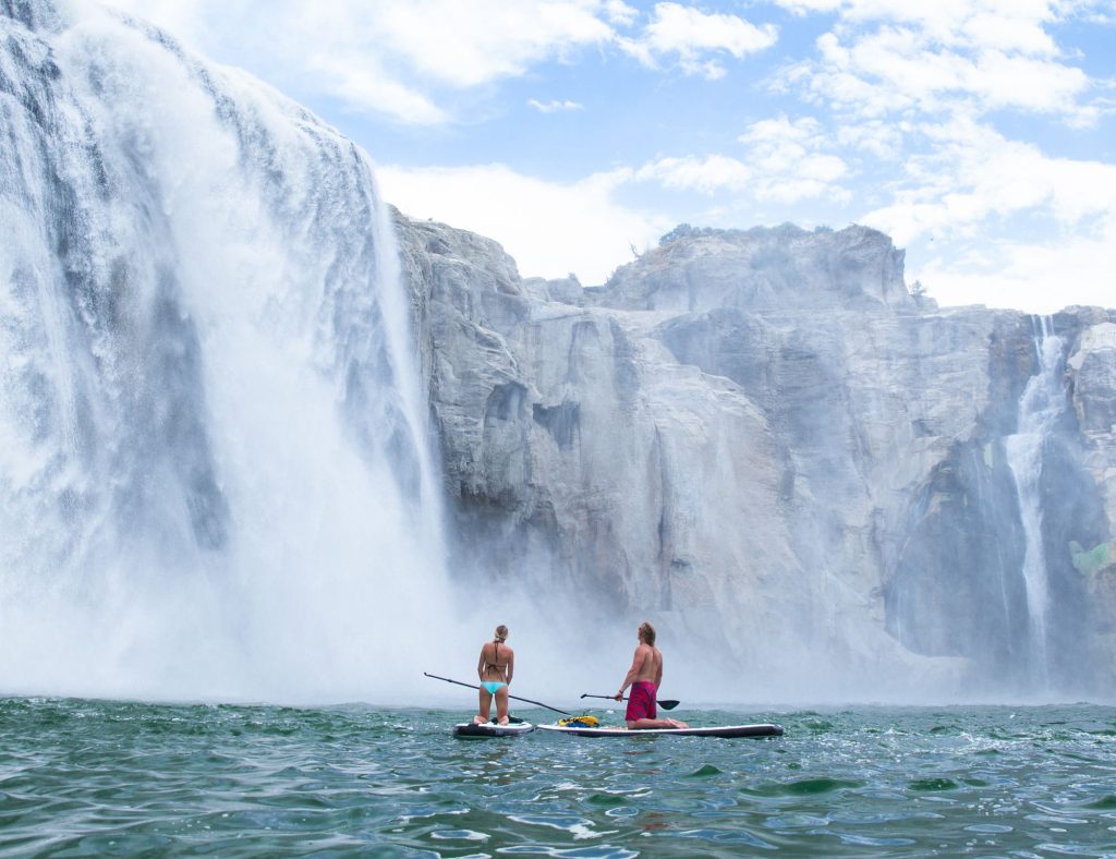 A couple kneels on stand-up paddleboards in front of a large, cascading waterfall.