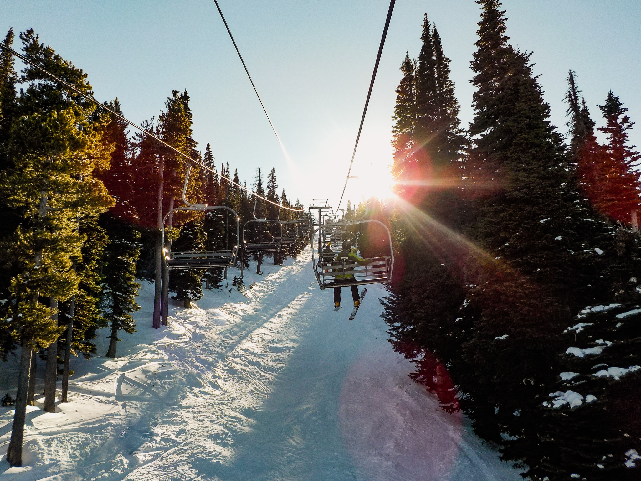 Two skiers ride on two lifts through evergreen trees during sunrise at Pomerelle Mountain Resort.