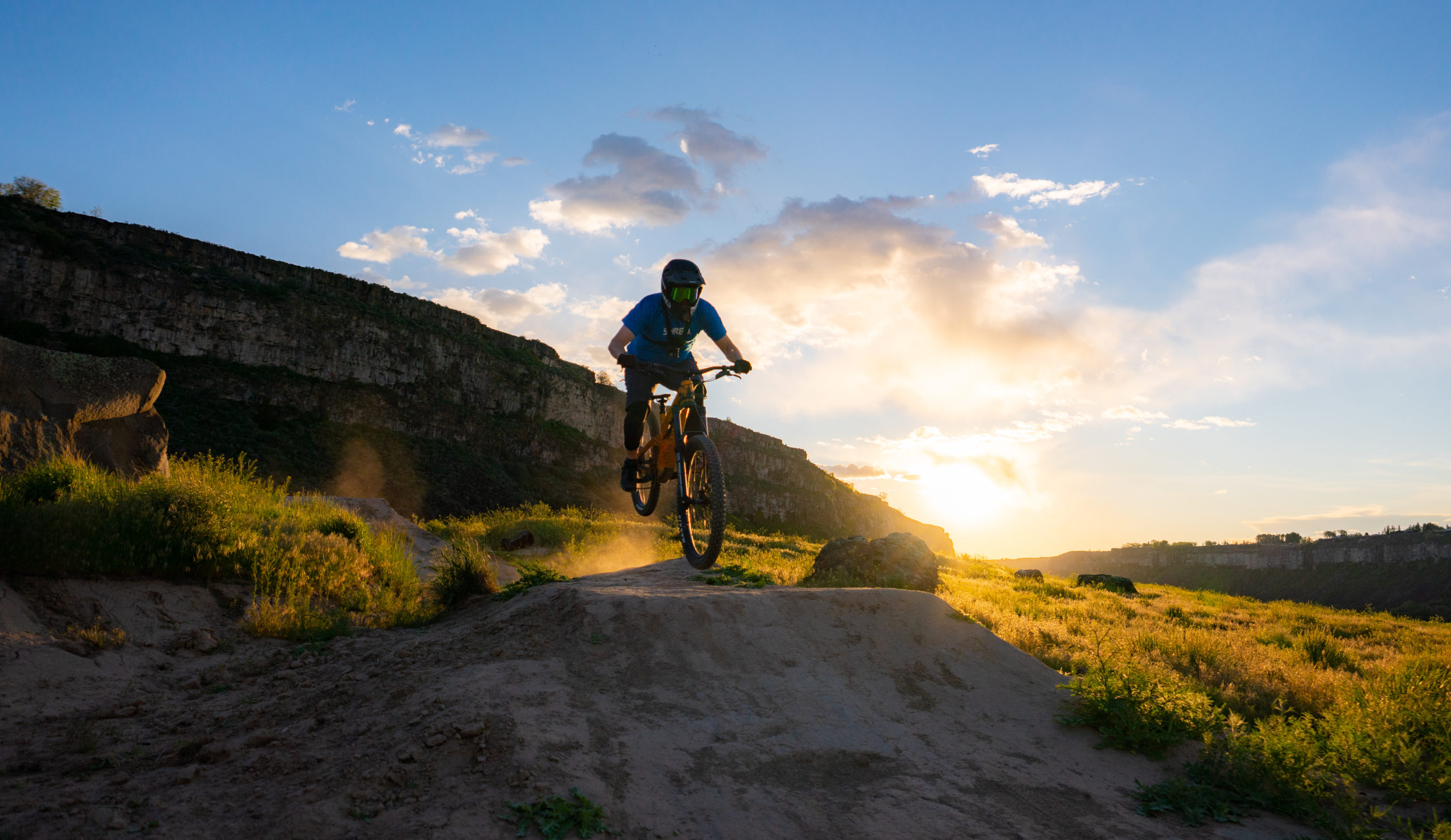 Man in helmet and glasses mountain biking at sunset, dust trailing across rugged terrain and yellow grass.