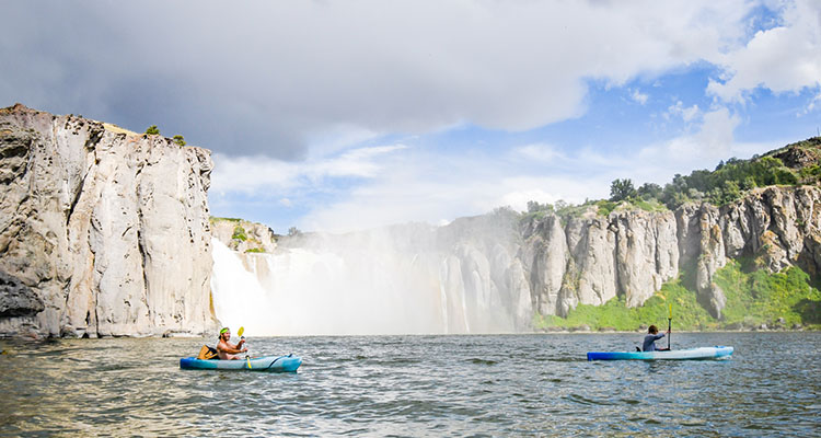Two people in blue kayaks paddle in front of Shoshone Falls, a large waterfall.