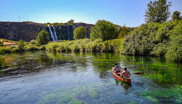 Two people in an orange canoe paddle on a river toward a waterfall at Ritter Island in southern Idaho.