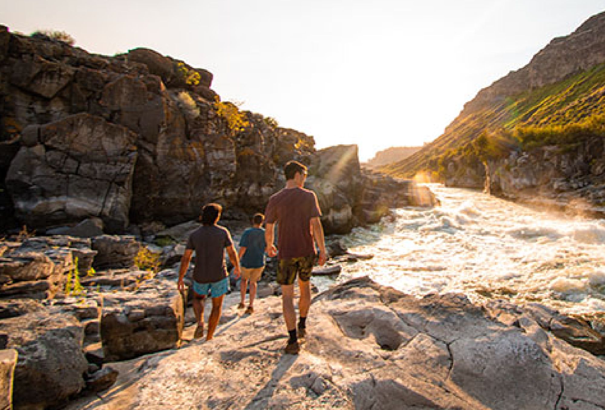 Three men walk across rugged terrain as the sun glows over crashing waters.