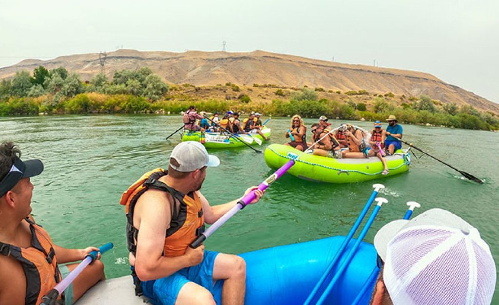 Three groups laugh and point their oars from inflatable rafts on the whitewater, with South Idaho landscapes in the background.