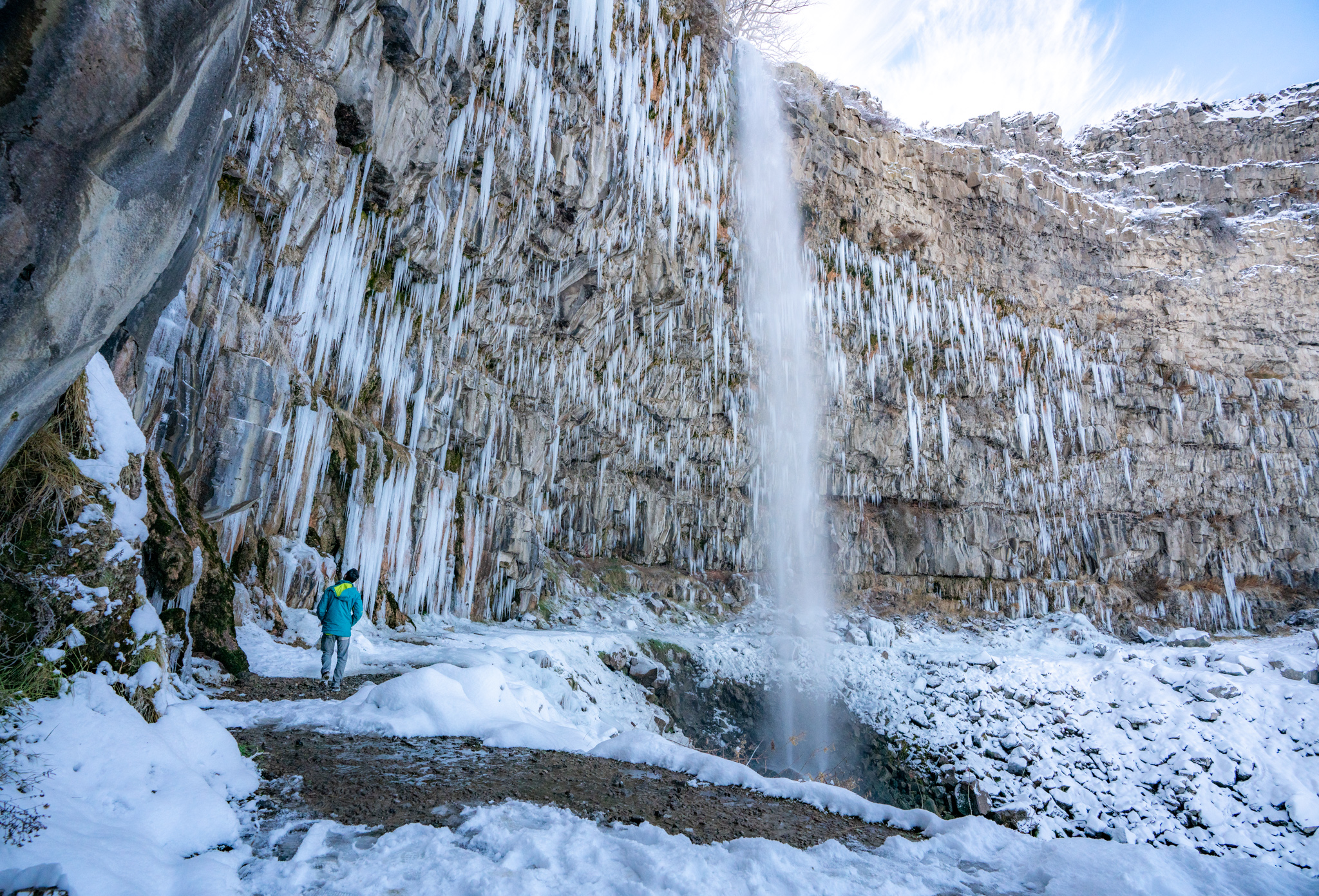 A person standing between a large rocky wall with frozen cascades of water and a waterfall flowing into a hole within a snowy landscape.