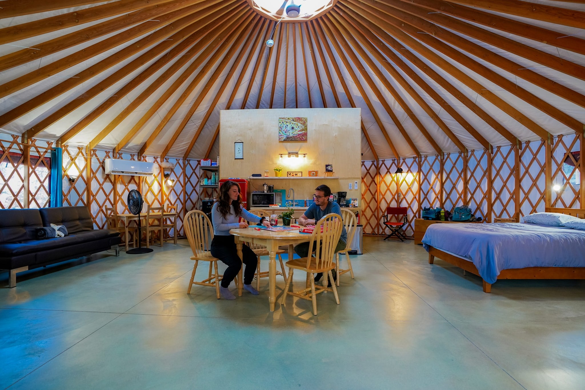Two people sit playing a board game at a wooden table in the middle of a large, well-decorated yurt.