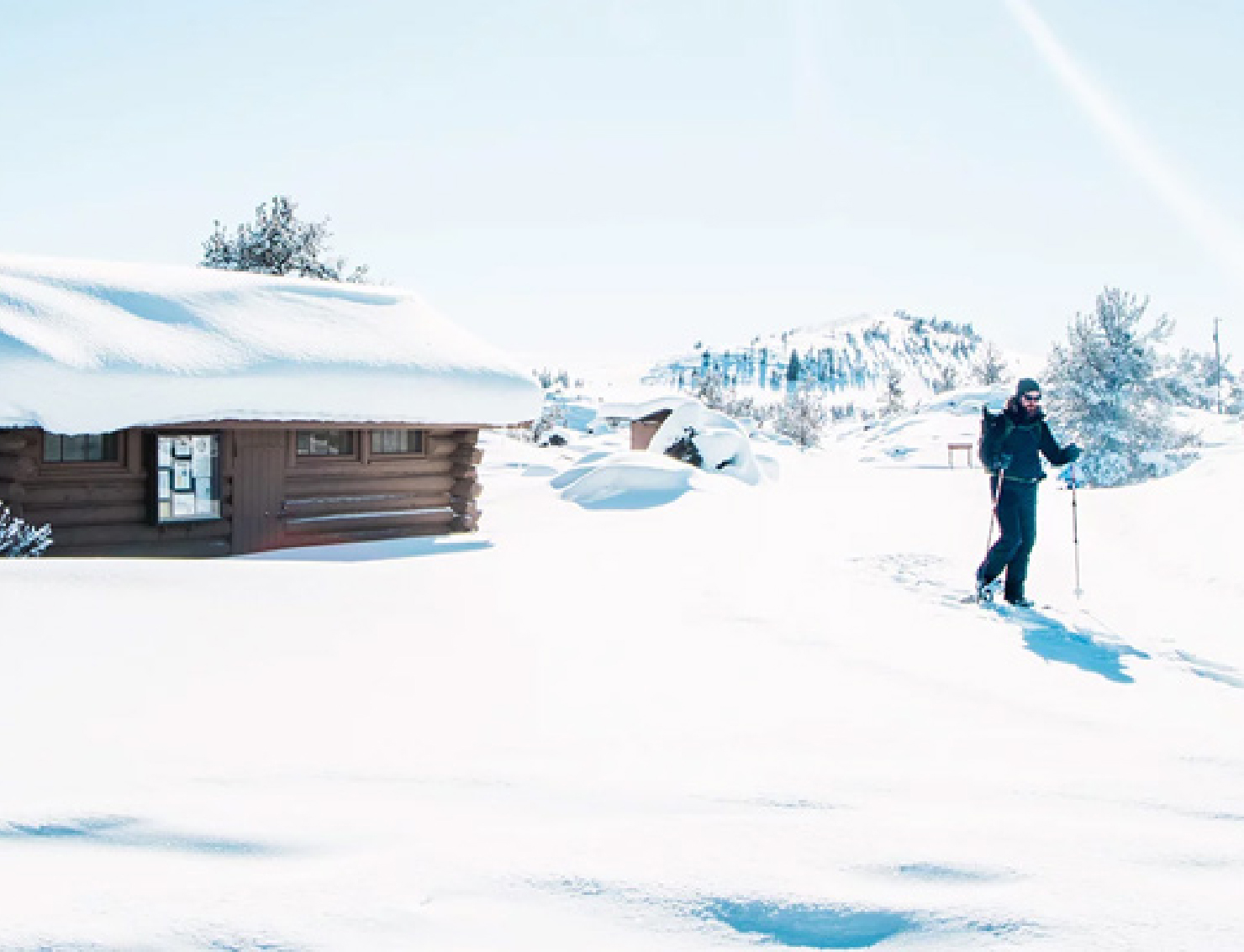 A person cross-country skiing across an open field of snow, and in the background, a small wooden lodge with a snow-covered roof.