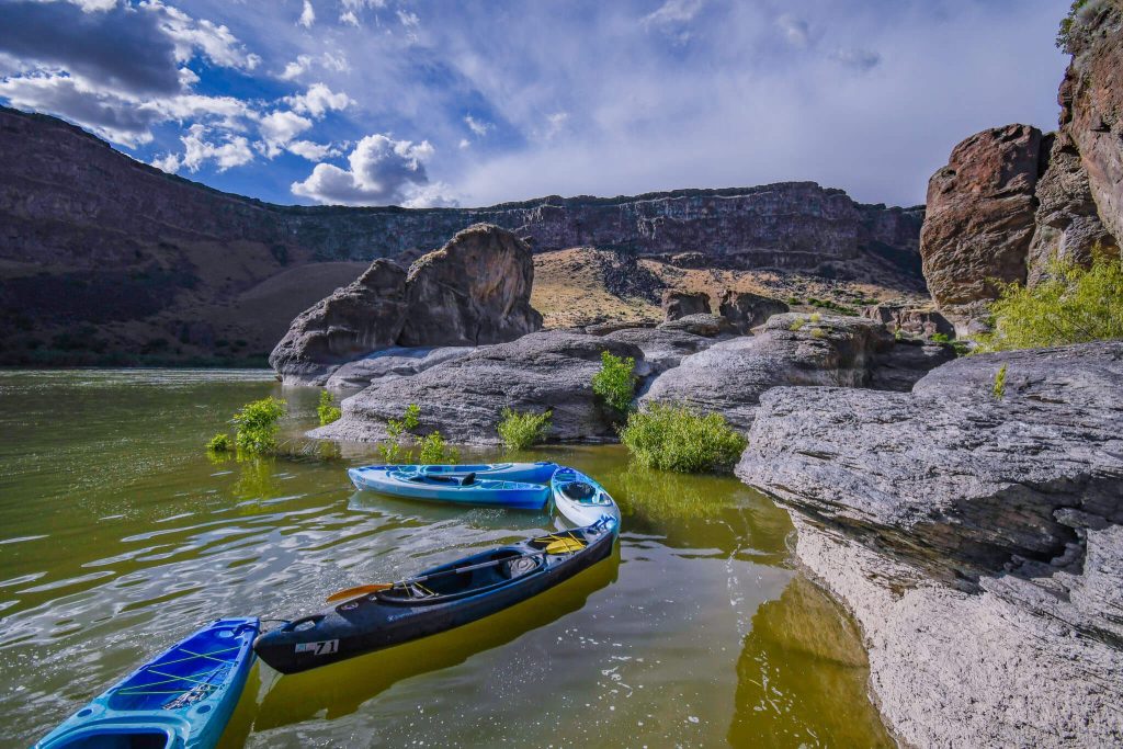 A group of blue kayaks in a body of water beside a group of large rocks surrounded by canyon walls.