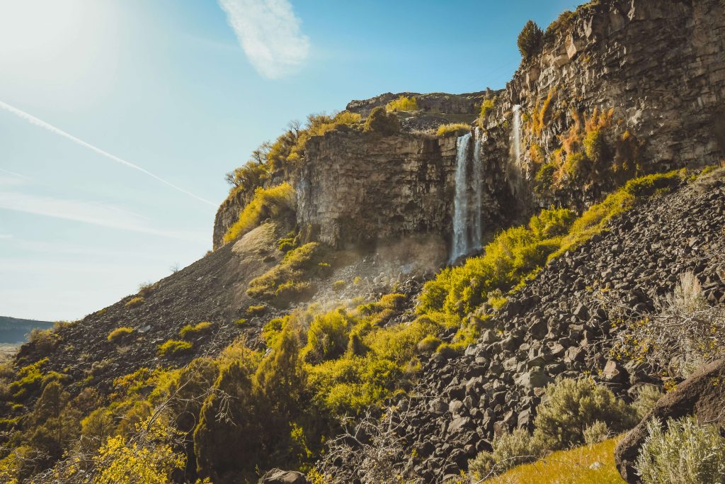 A waterfall cascading from the side of a cliff surrounded by brush and rocky landscapes.