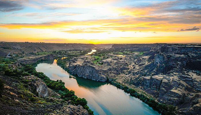 A view of the Snake River at sunset, lined with rocky landscapes and trees.