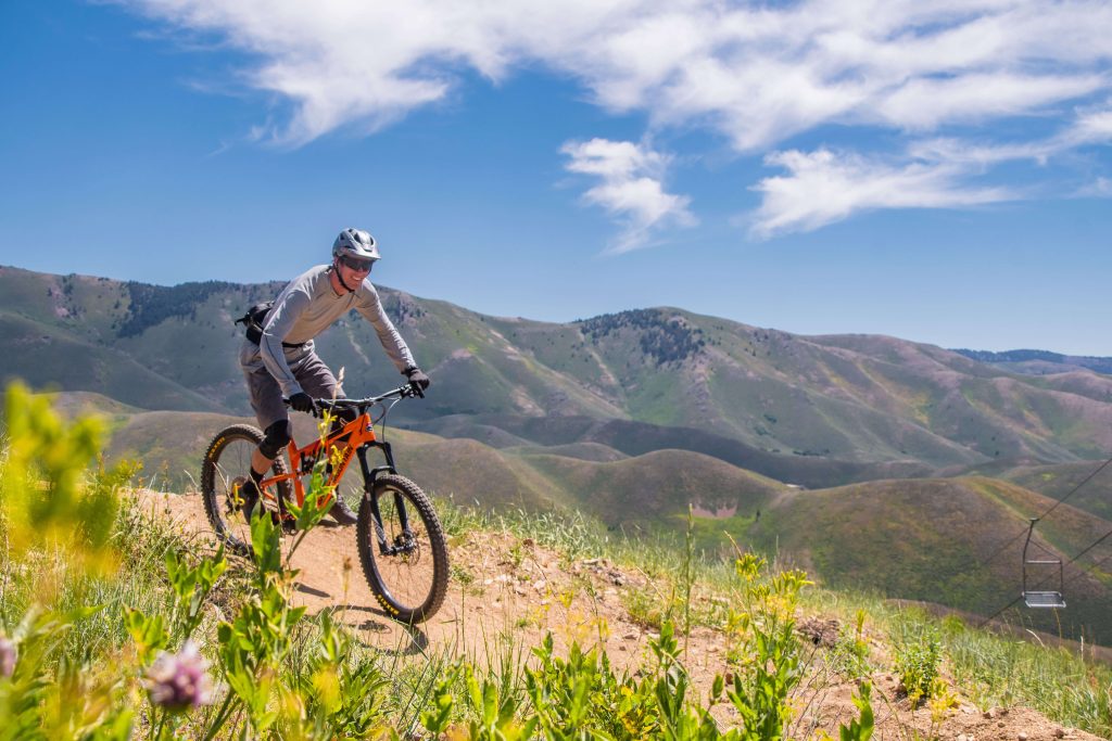 A person riding a mountain bike across a trail lined with tall grass at Soldier Mountain Resort, and in the background, a series of mountains.