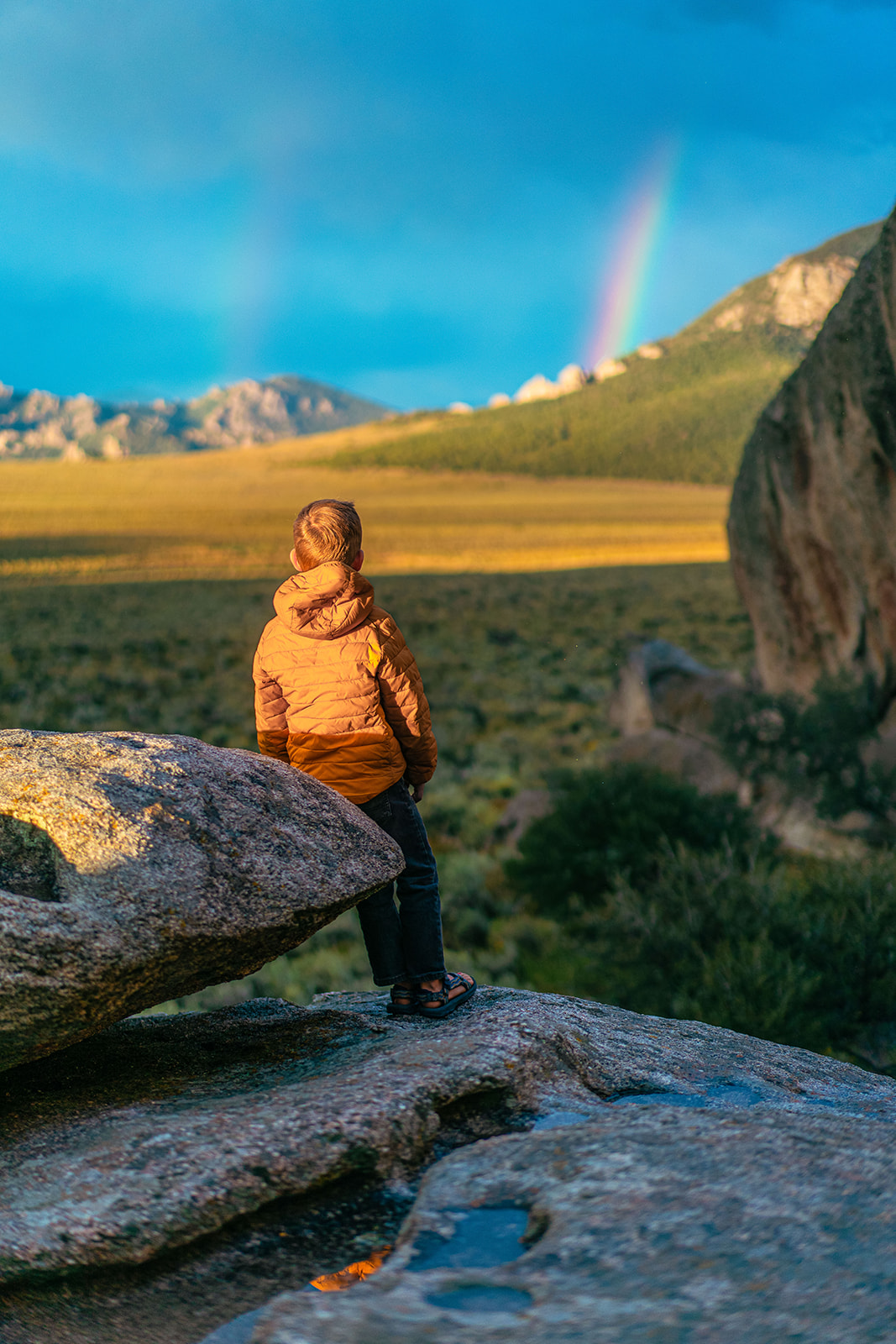 A child standing on a large rock and leaning on another large rock, looking at a vast green landscape and a double rainbow in the distance.