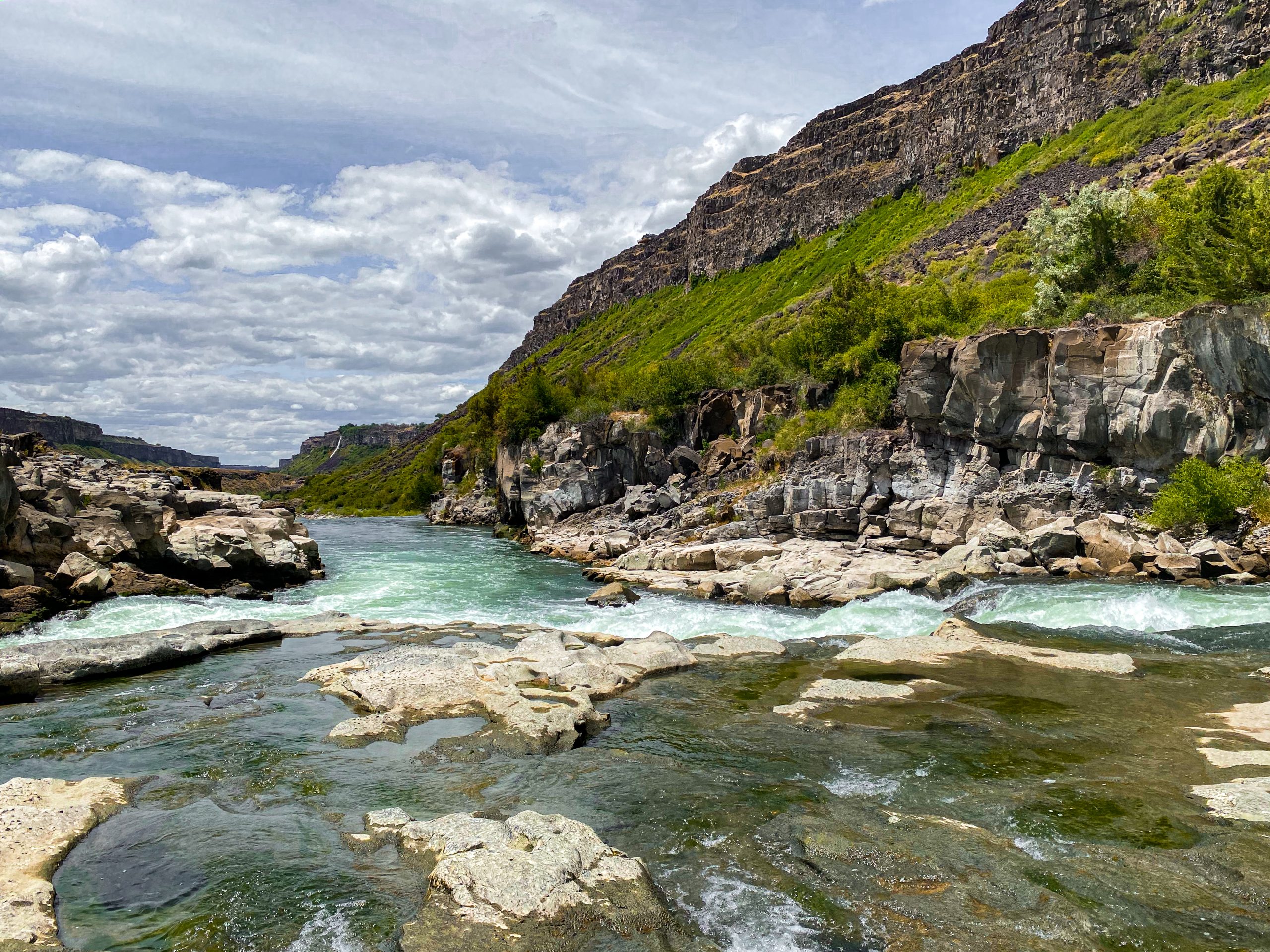 Large rocks in a river lined with rocky terrain and green bushes within a canyon.