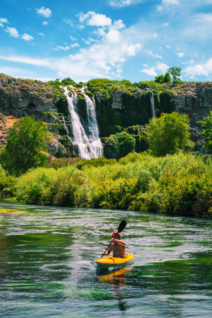 A person kayaking on a body of water at Thousand Springs State Park, and in the background, tall trees, bushes and a waterfall spilling over a rocky wall.