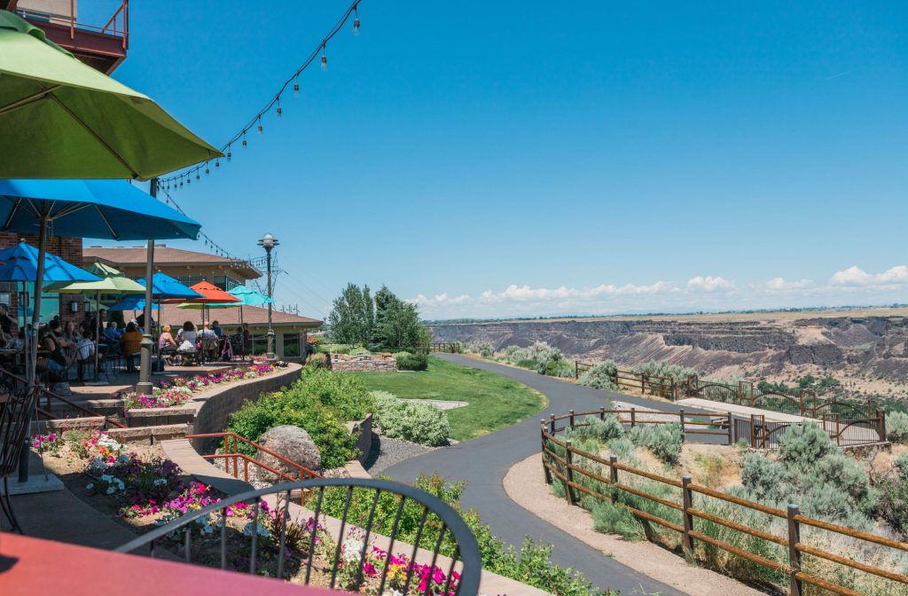 A vibrant patio filled with bright flowers and colorful umbrellas overlooks a winding path through the sprawling green landscape of Southern Idaho.