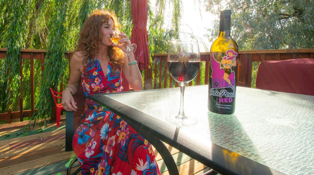 A girl in a vibrant floral dress sips Ida Peach Red wine, with leafy trees peeking through a wooden fence.