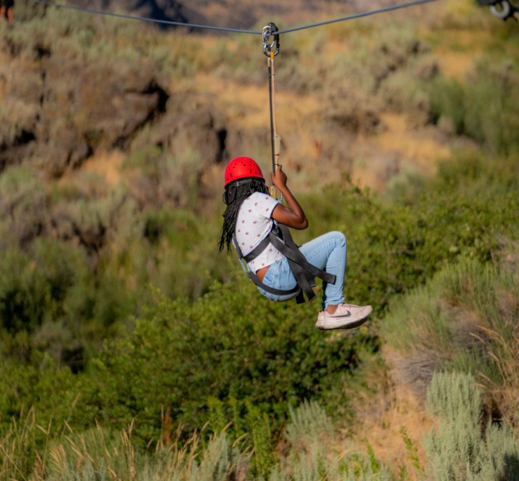 A girl wearing a red helmet soars on a zipline above lush green terrain.