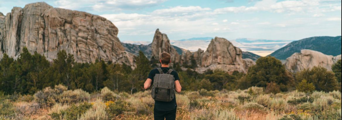 Man with backpack standing in wild grasses, overlooking rugged Southern Idaho mountains under a bright, cloud-filled sky.