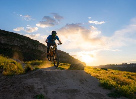Man in helmet and glasses mountain biking at sunset, dust trailing across rugged terrain and yellow grass.