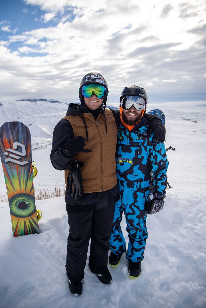 Two snowboarders with arms around each other and big grins, wearing helmets and goggles, with a snowboard behind them on the snowy slopes of Soldier Mountain beneath a bright, cloudy sky.