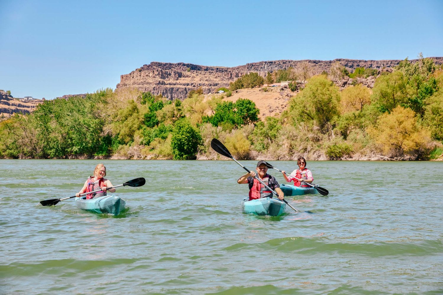 Three kayakers on the Snake River at Centennial Waterfront Park in Twin Falls, Idaho with the Perrine Bridge in the background.